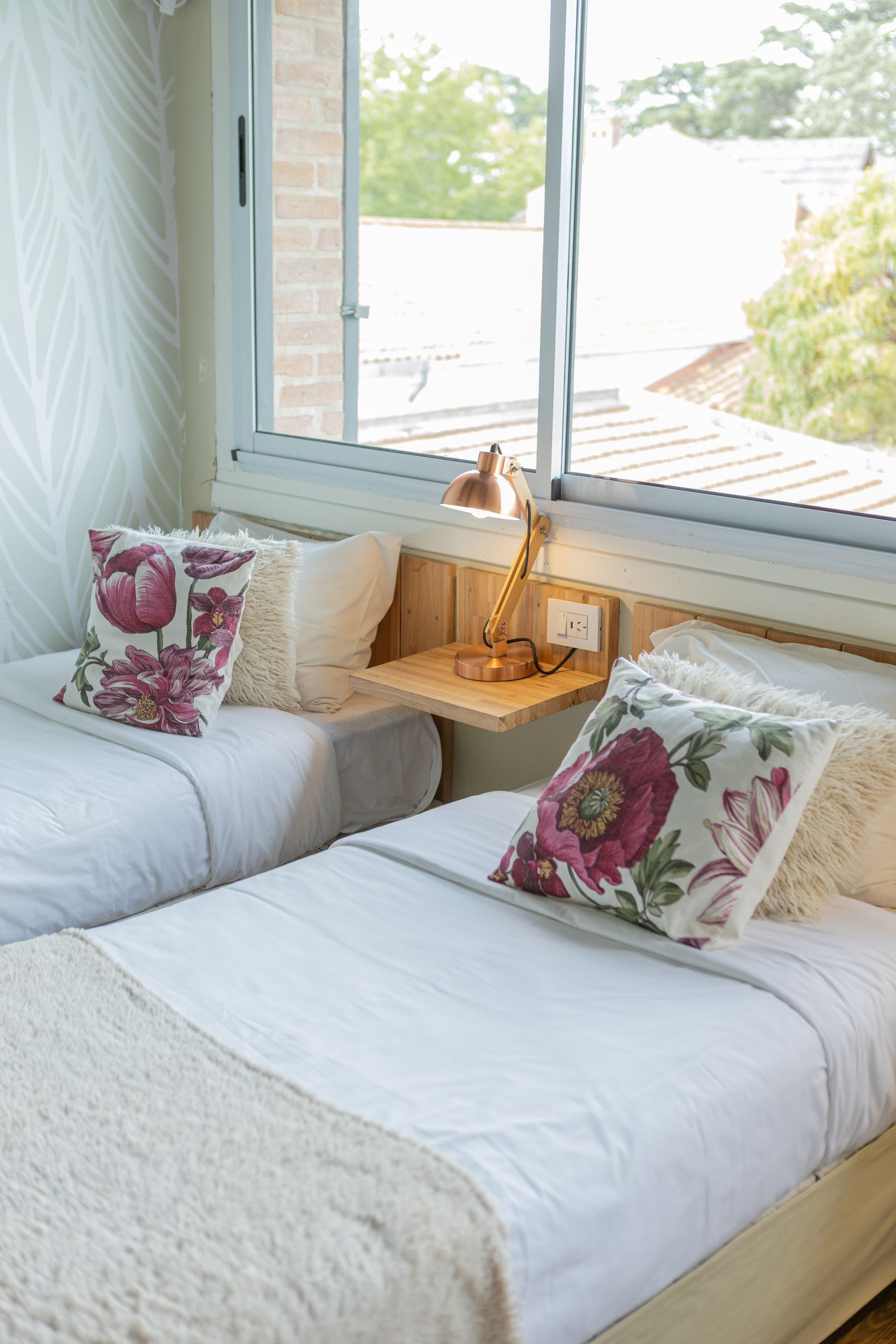 Dos camas individuales bien hechas junto a una ventana luminosa con cojines estampados con flores y una pequeña lámpara de noche.