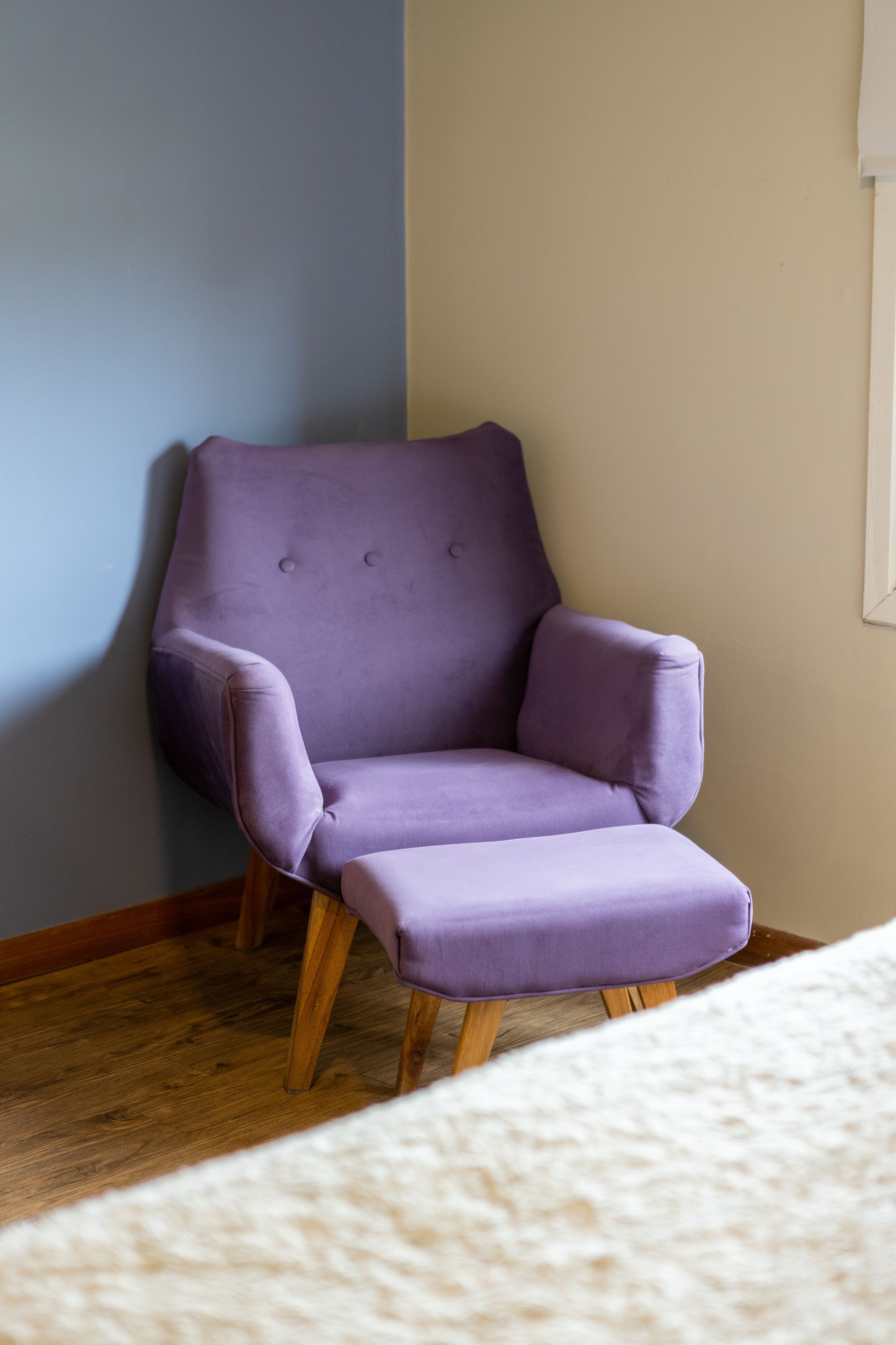 Sillón lavanda con otomana en un rincón junto a una ventana, sobre un suelo de madera.
