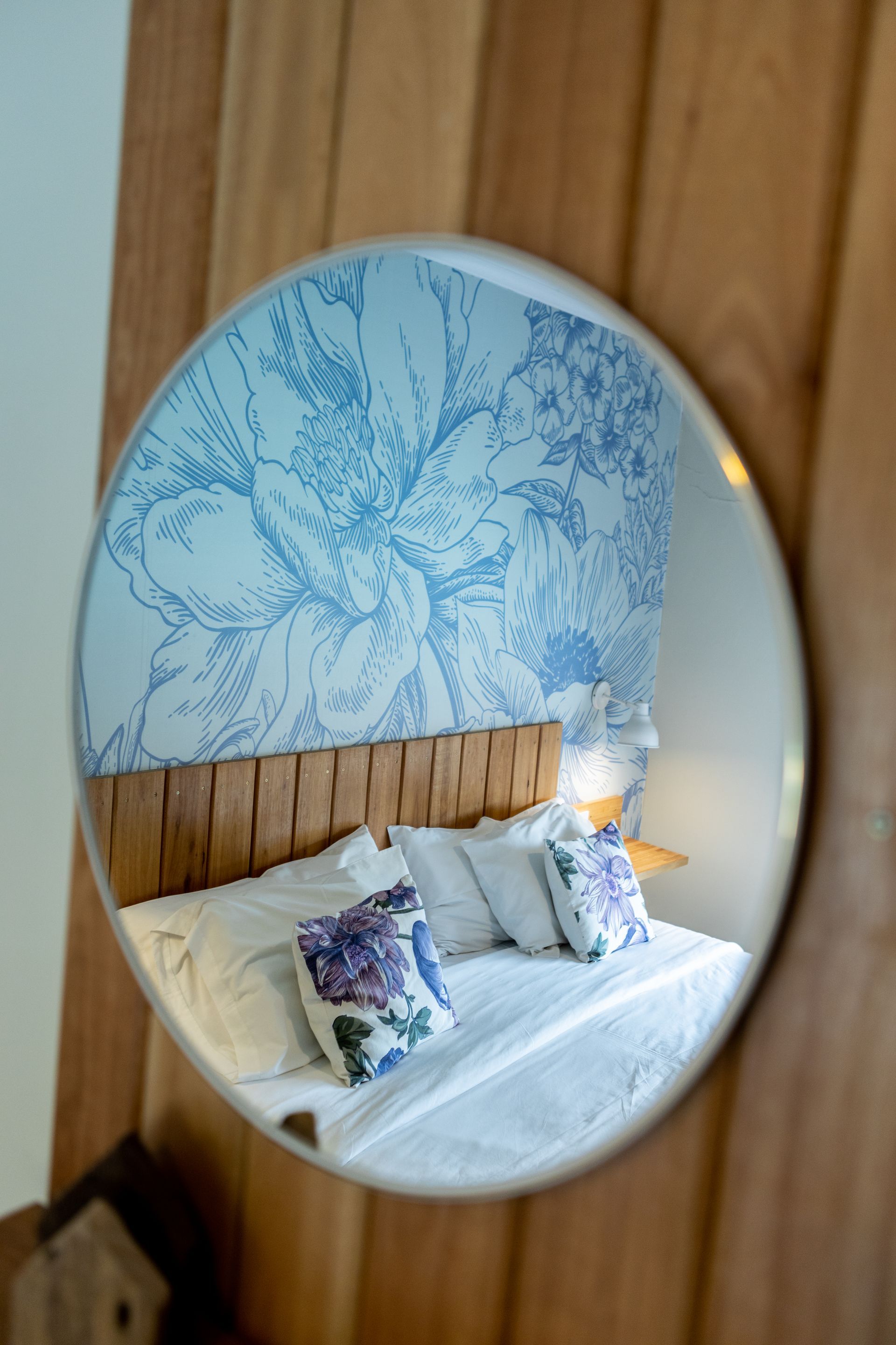 Espejo ovalado que refleja una cama acogedora con papel tapiz botánico azul y ropa de cama blanca.