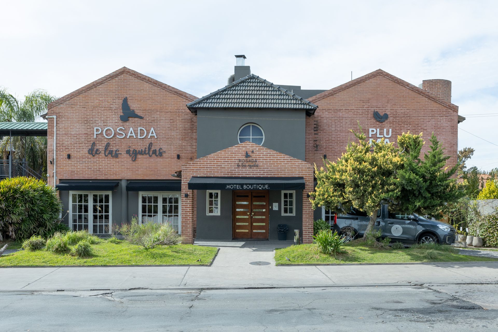 Local comercial de ladrillo con entrada cubierta, vegetación y un coche aparcado a la derecha.