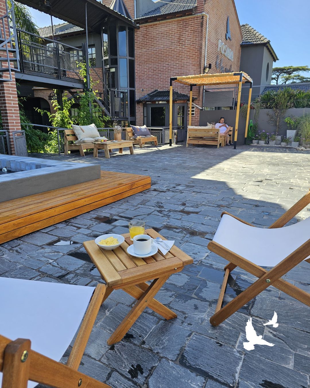 Patio exterior con tumbonas de madera, una mesita y una casa de ladrillo al fondo.