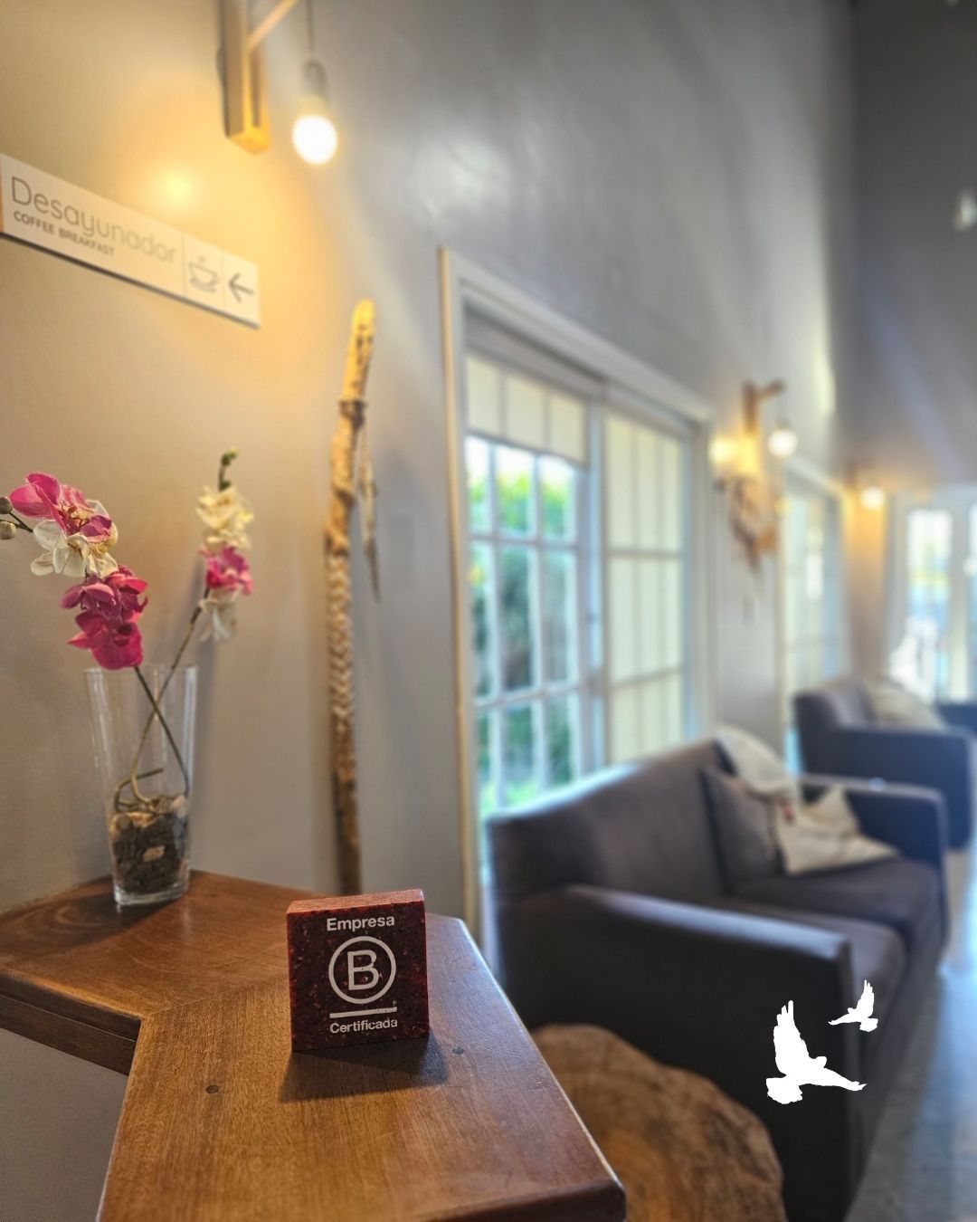 Interior acogedor de una cafetería con una mesa de madera, flores, luces de pared cálidas y sillones junto a la ventana.