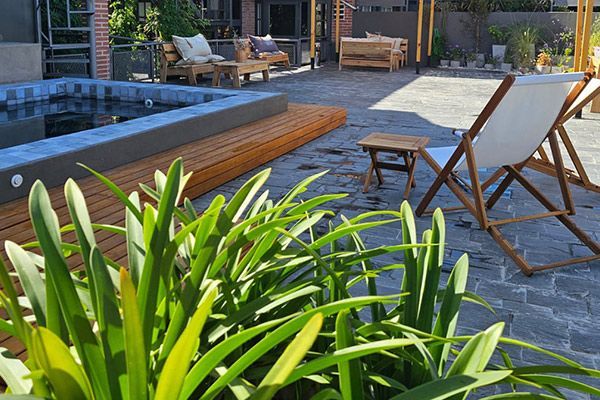 Piscina moderna en el patio trasero con tumbonas, terraza de madera y exuberantes plantas verdes en primer plano.