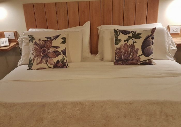 Cama bien hecha con sábanas blancas, dos almohadas con estampado floral y cabecero de madera en una habitación de hotel.