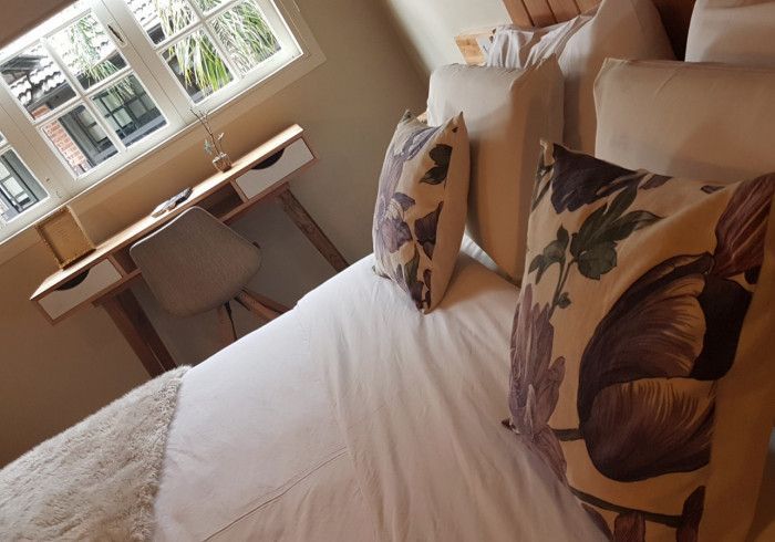 Habitación luminosa con una cama bien hecha, cojines estampados con flores y un escritorio junto a la ventana.