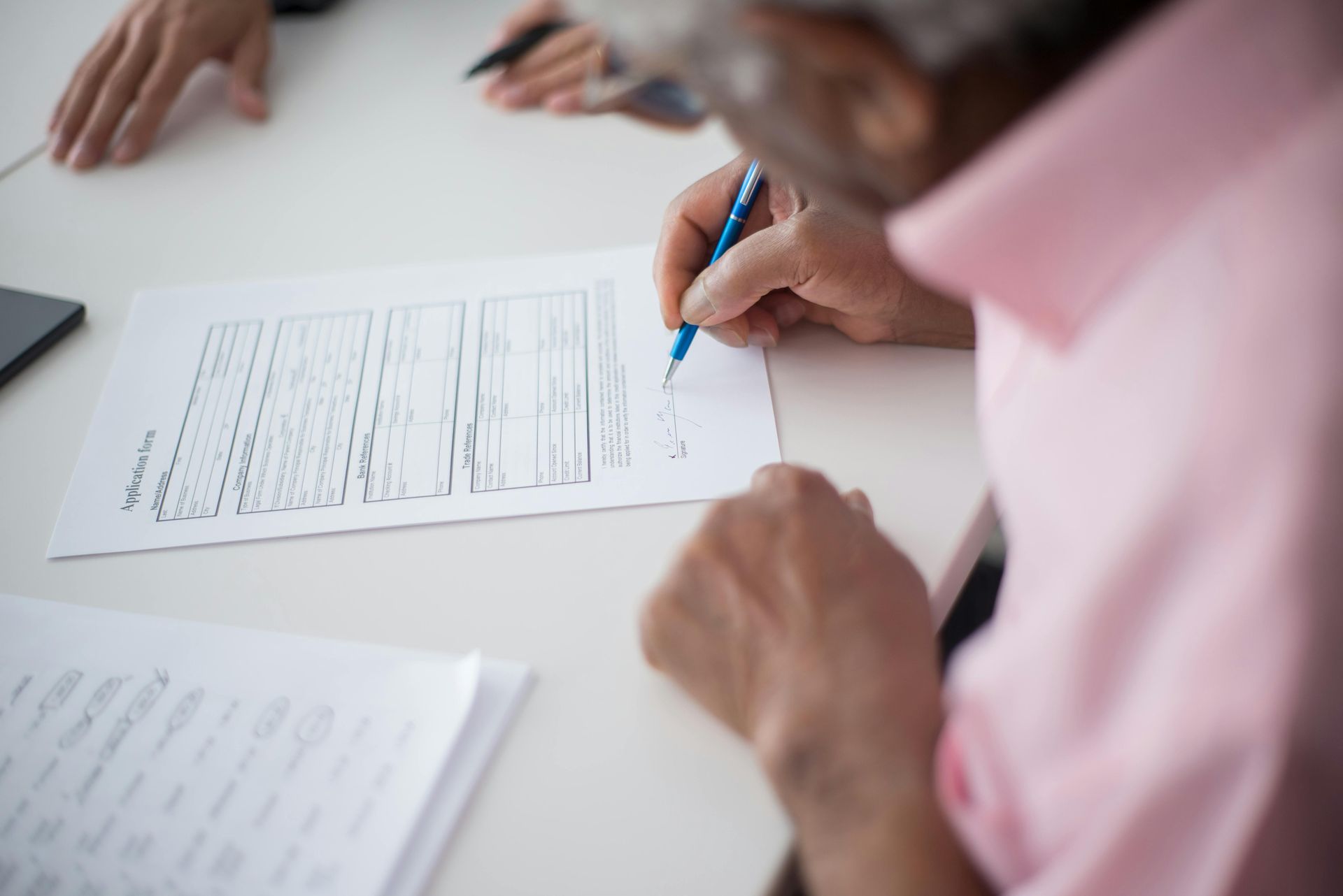 Person signing a document at a table with others; a blue pen is used.