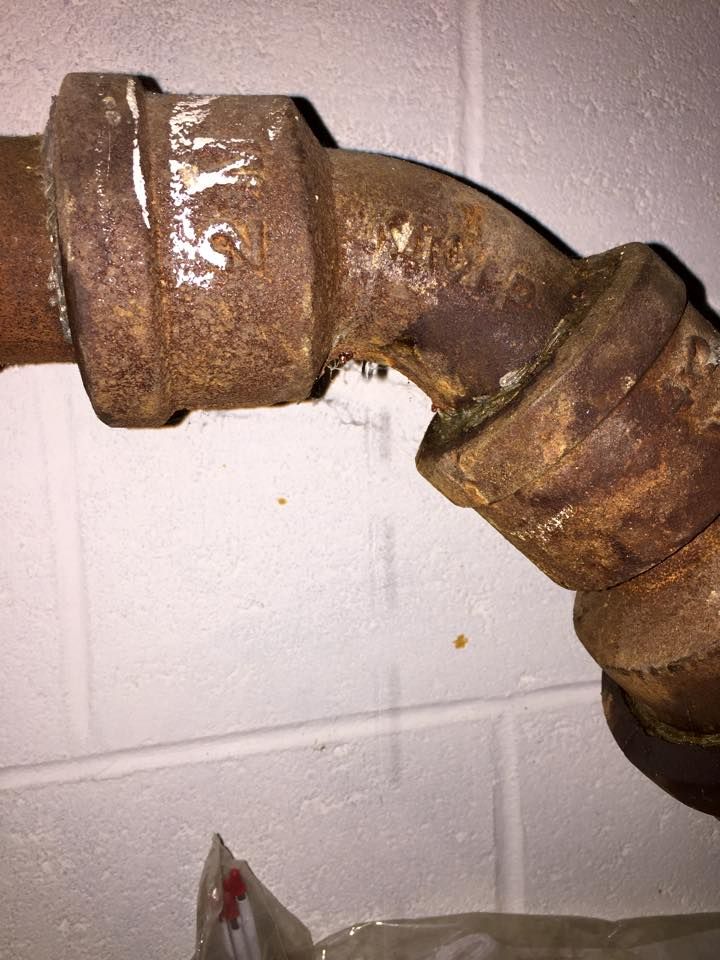 Rusty metal pipe elbow joint on a white wall, showing signs of leakage.