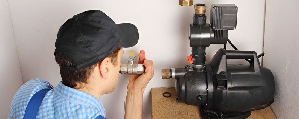 Plumber examining a water pump in a confined space. He wears a cap and blue shirt.