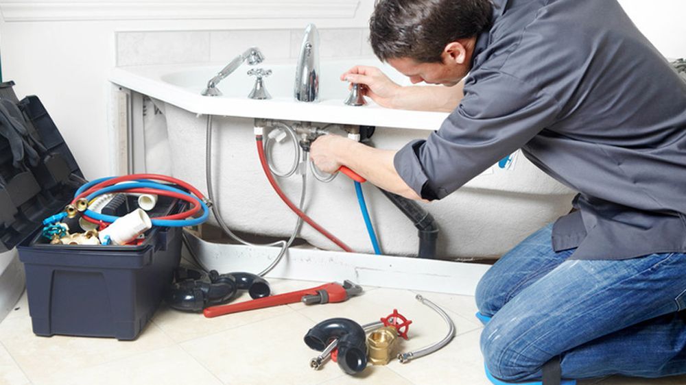 Plumber kneeling by a bathtub, working on pipes. Tools and toolbox nearby; focus on plumbing.