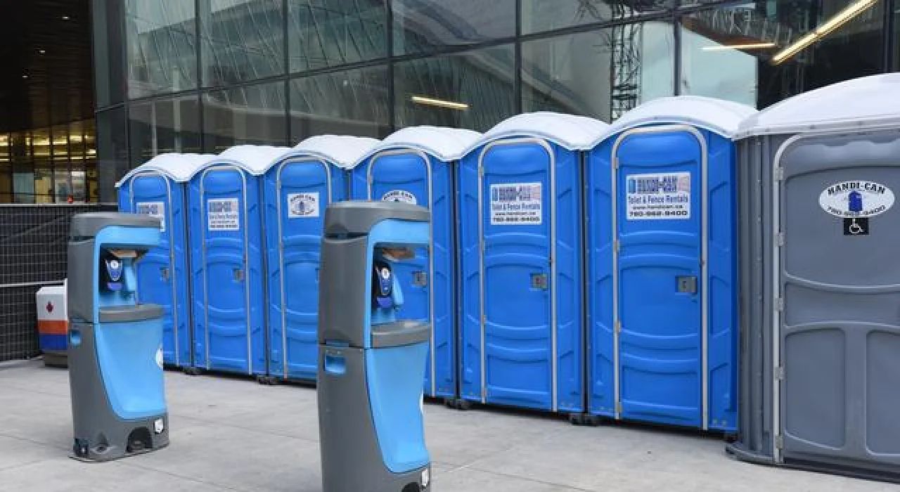 Row of blue portable toilets outside a glass building, with two blue hand sanitizer stations in front