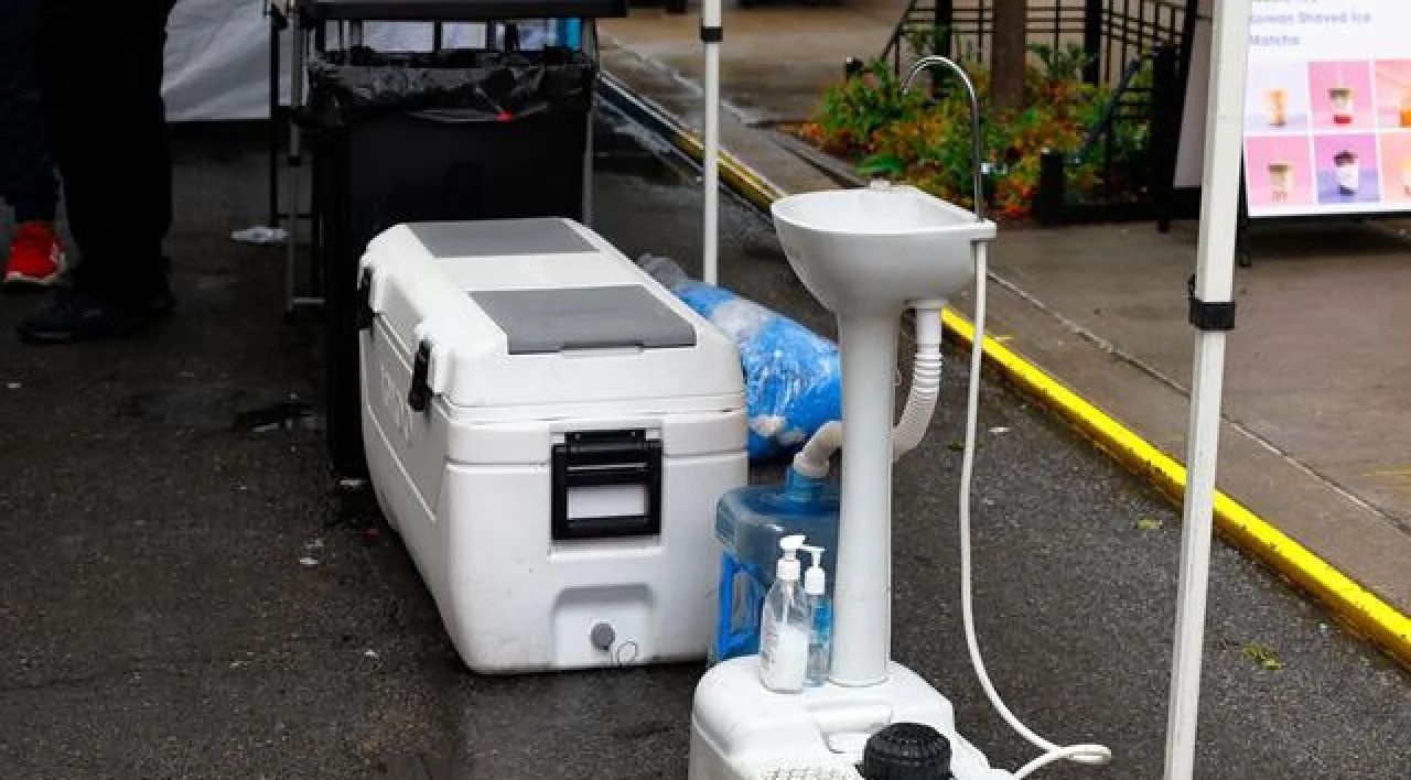 White cooler and handwashing station on wet pavement beside a curb and plants