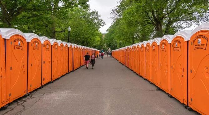 Orange portable toilets line both sides of a tree-lined park path with a few people walking ahead.