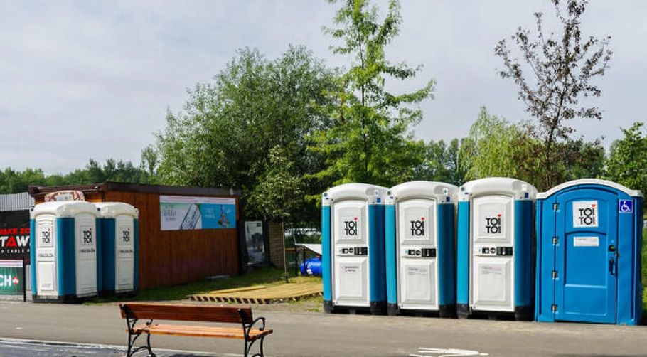 Row of blue-and-white portable toilets beside a brick building in a parking lot with a bench in front
