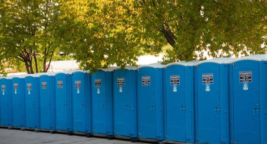 Row of blue portable toilets lined up outdoors beneath green trees