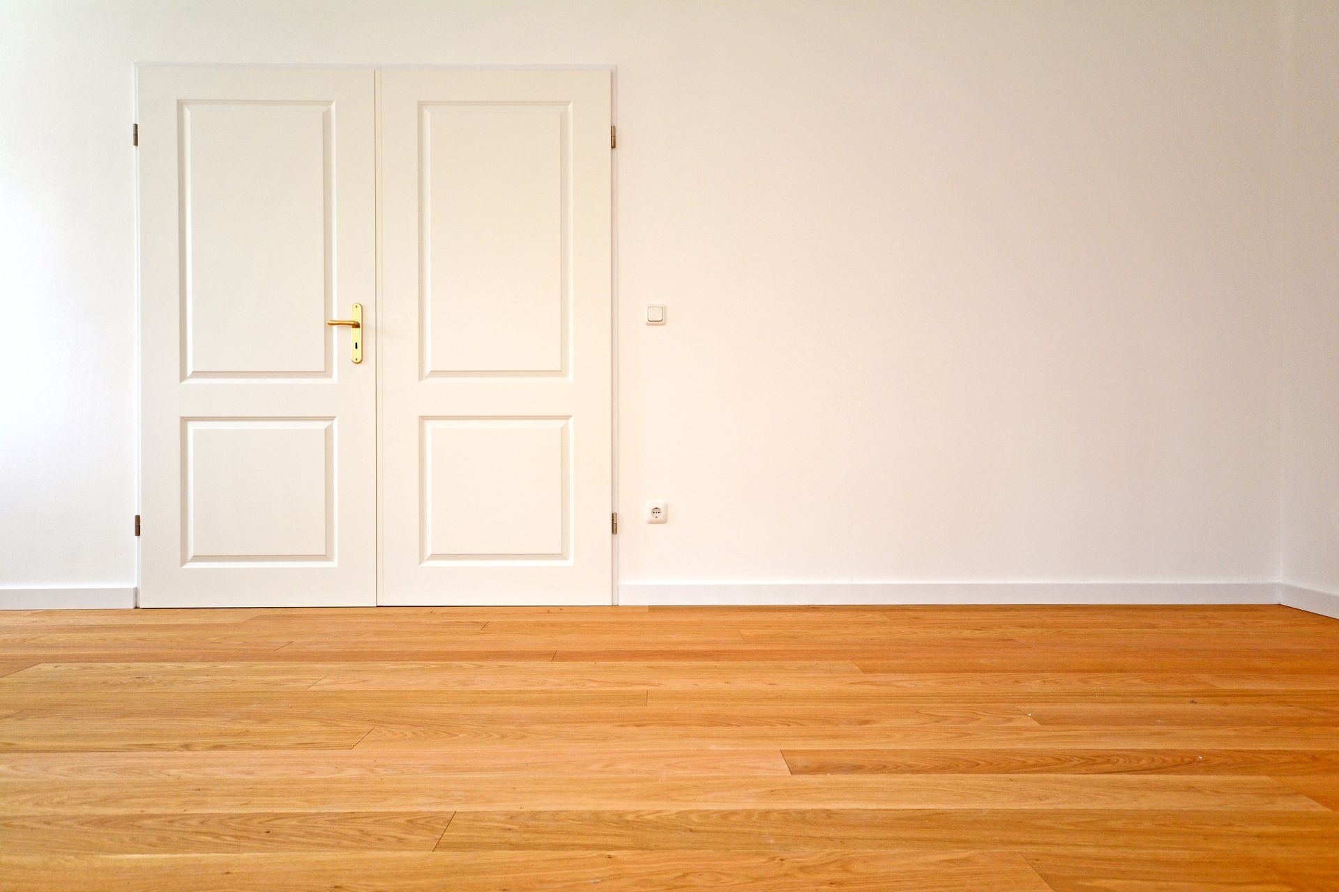 Ein Paar weiße Doppeltüren vor einer schlichten weißen Wand über hellem Holzboden in einem hellen, leeren Raum.