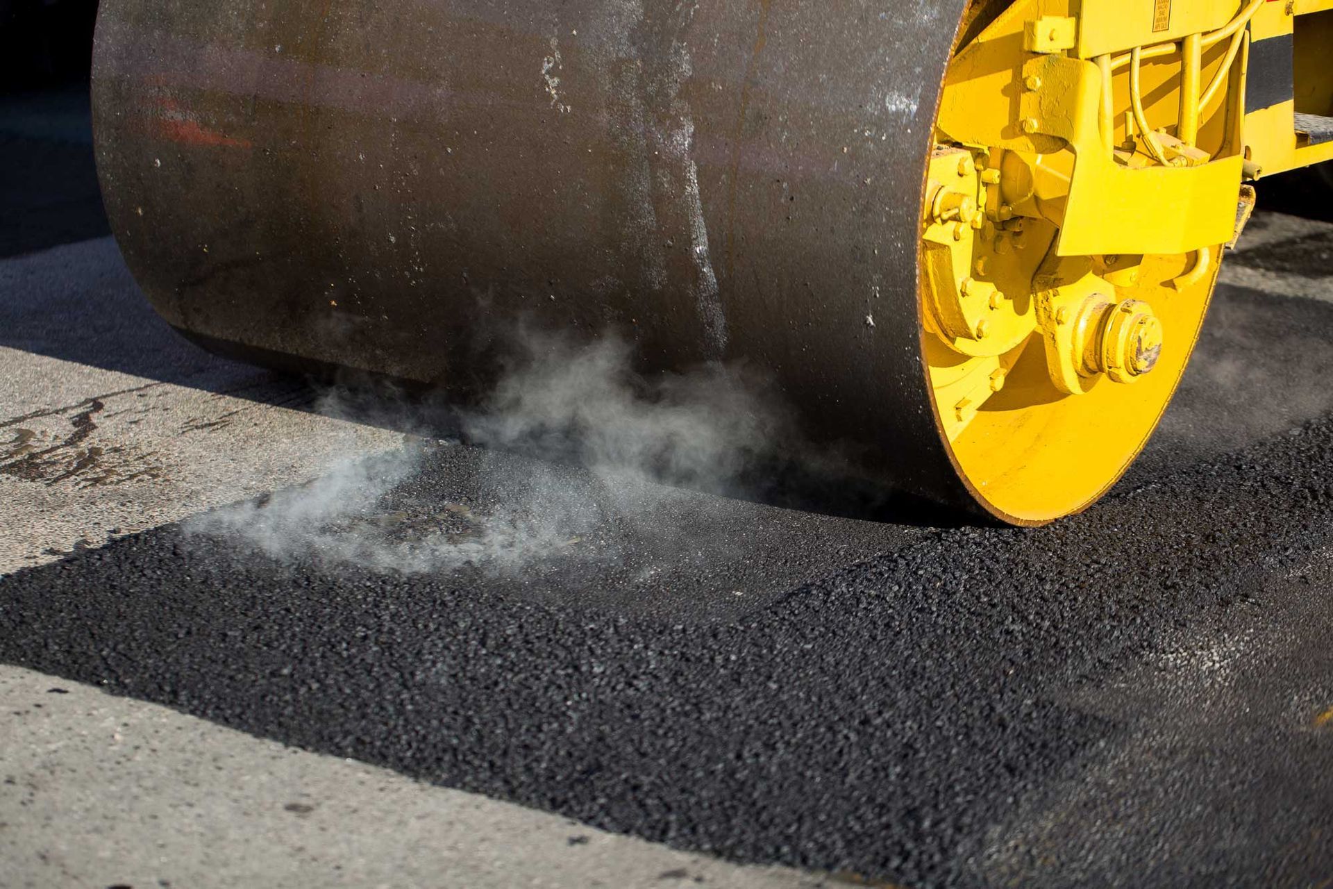 Steam rises as a heavy roller presses fresh asphalt during road paving work.