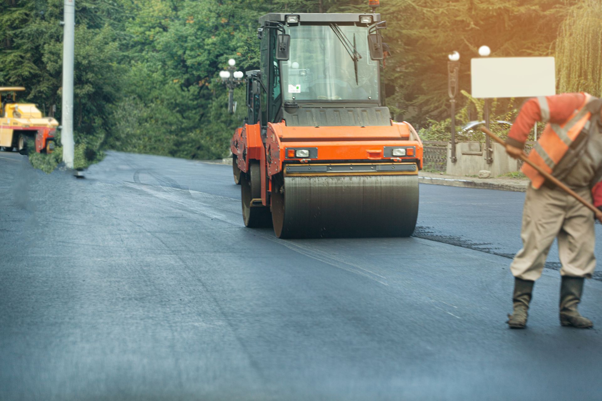 Road roller paving fresh asphalt as worker smooths road with tool nearby.