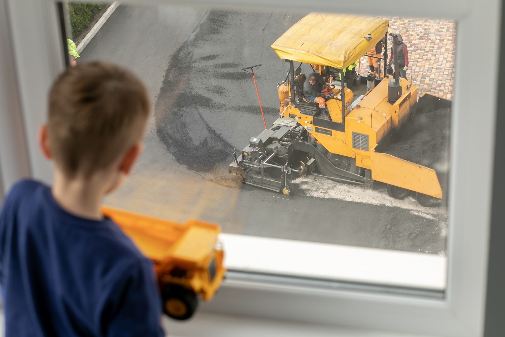 Boy looks out of the window as pavers work below. They lay asphalt on the driveway.