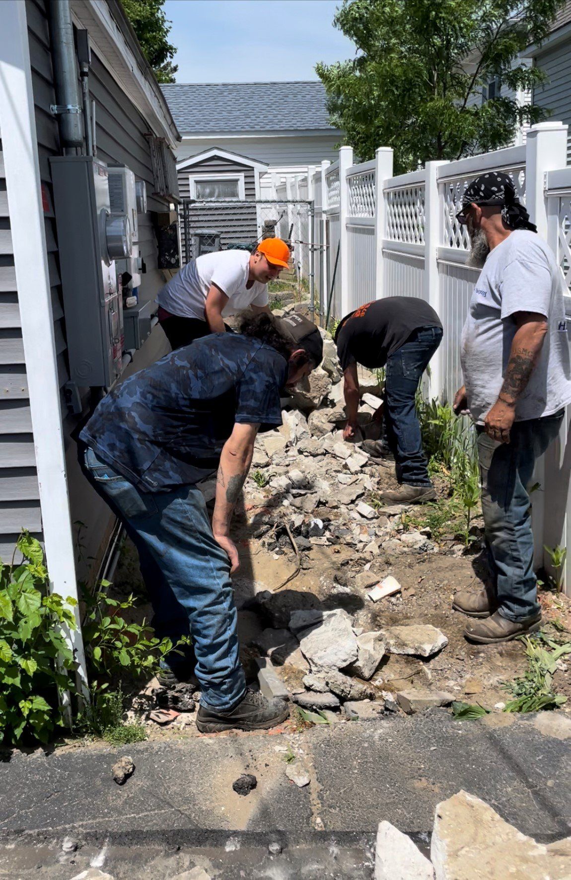 Four people in casual work attire clearing broken concrete and debris from a narrow side yard between two buildings.