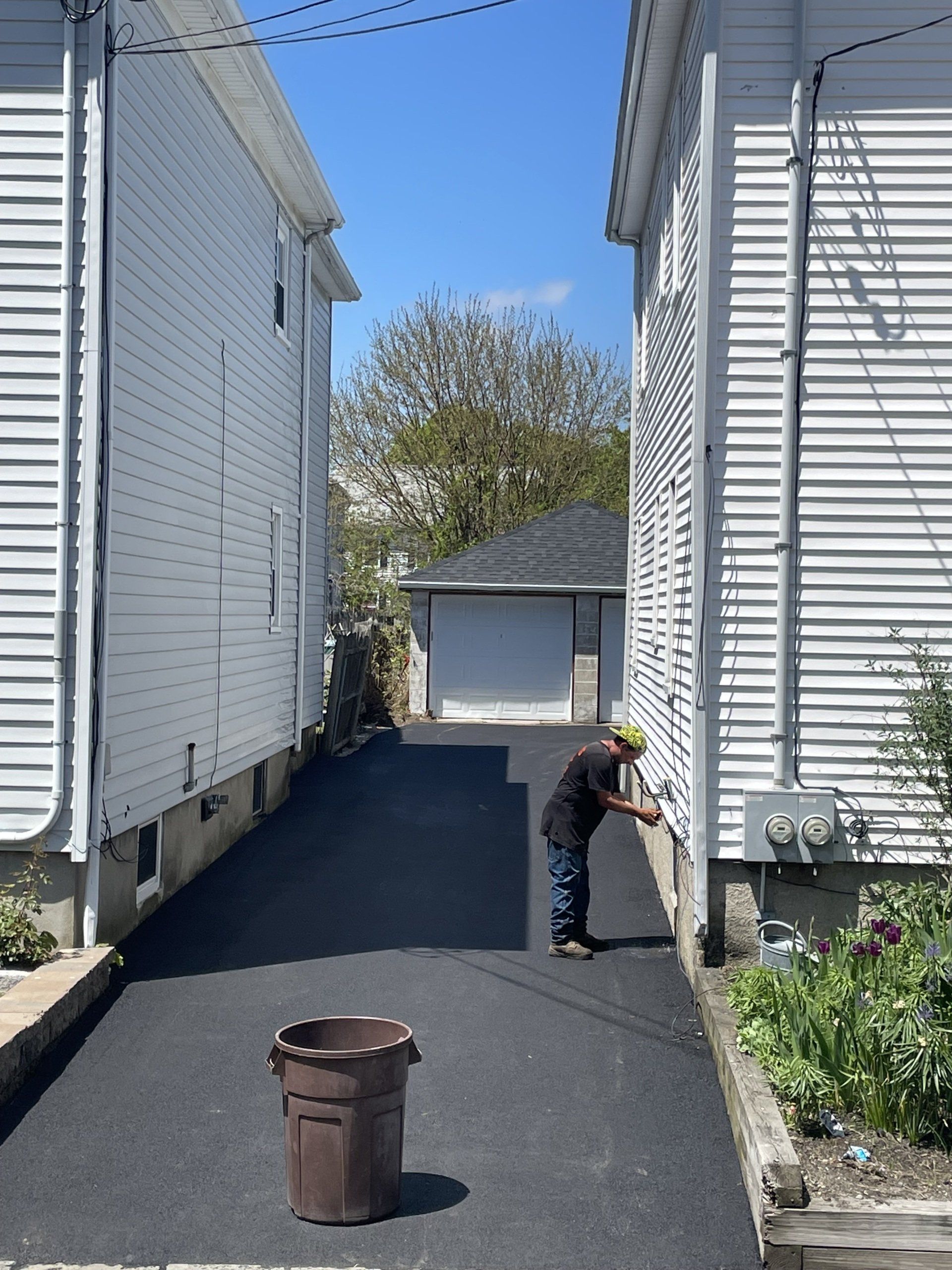 A person in a dark shirt and jeans works near the siding of a white house beside a newly paved driveway with a trash bin.
