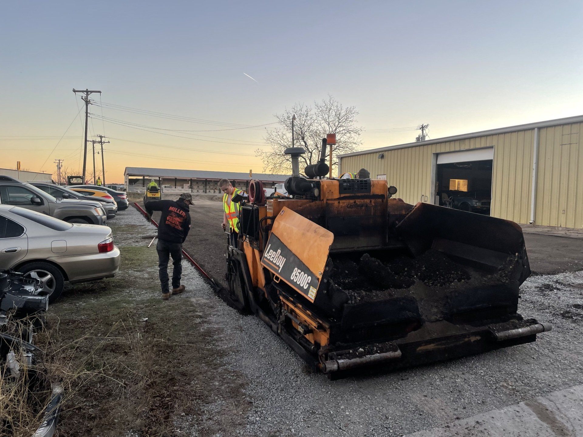 Construction crew operating a yellow asphalt paving machine in a gravel lot next to a warehouse building at sunset.