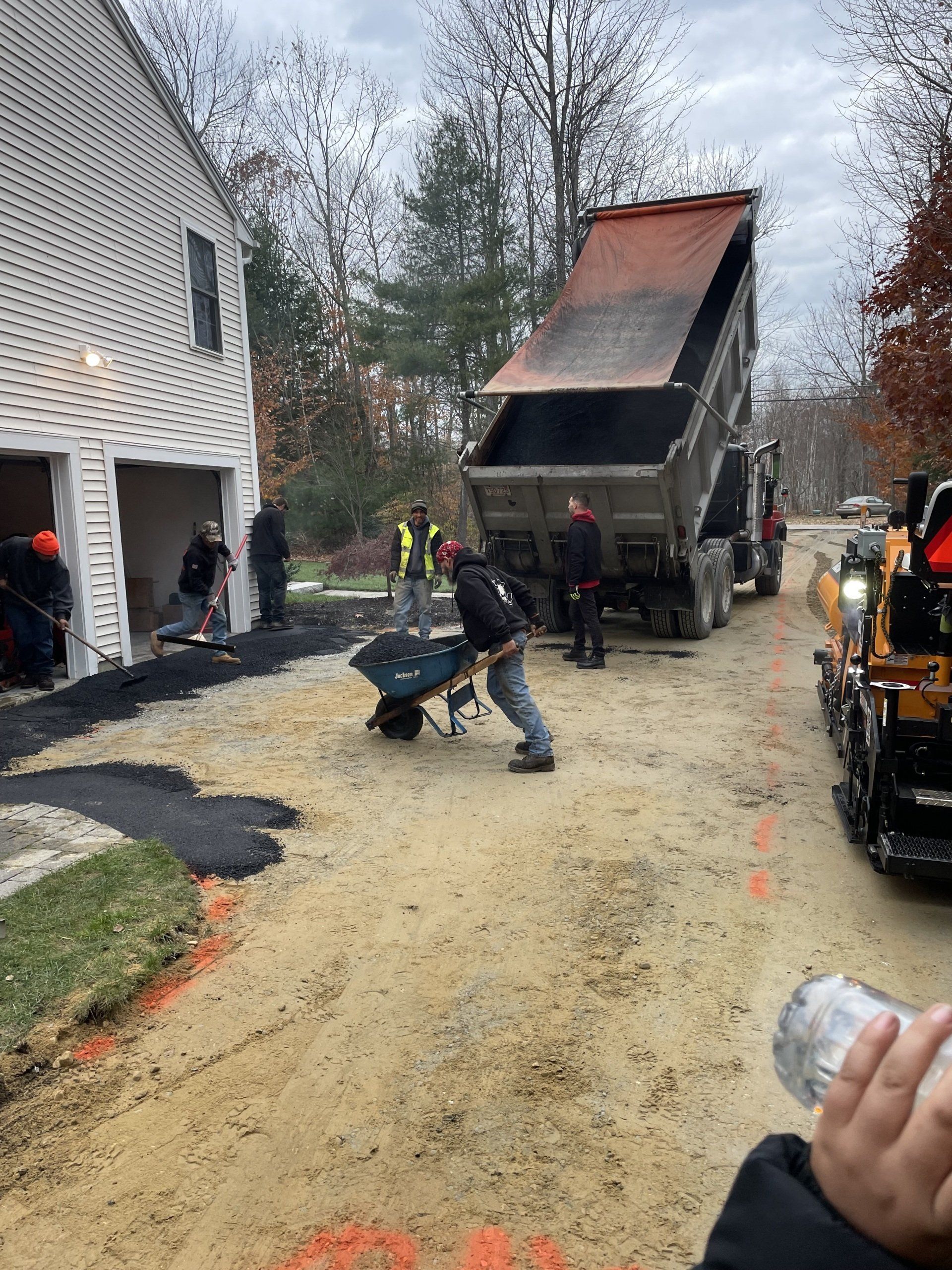 A crew works on a driveway project, with a dump truck and a worker pushing a wheelbarrow near a house under construction.