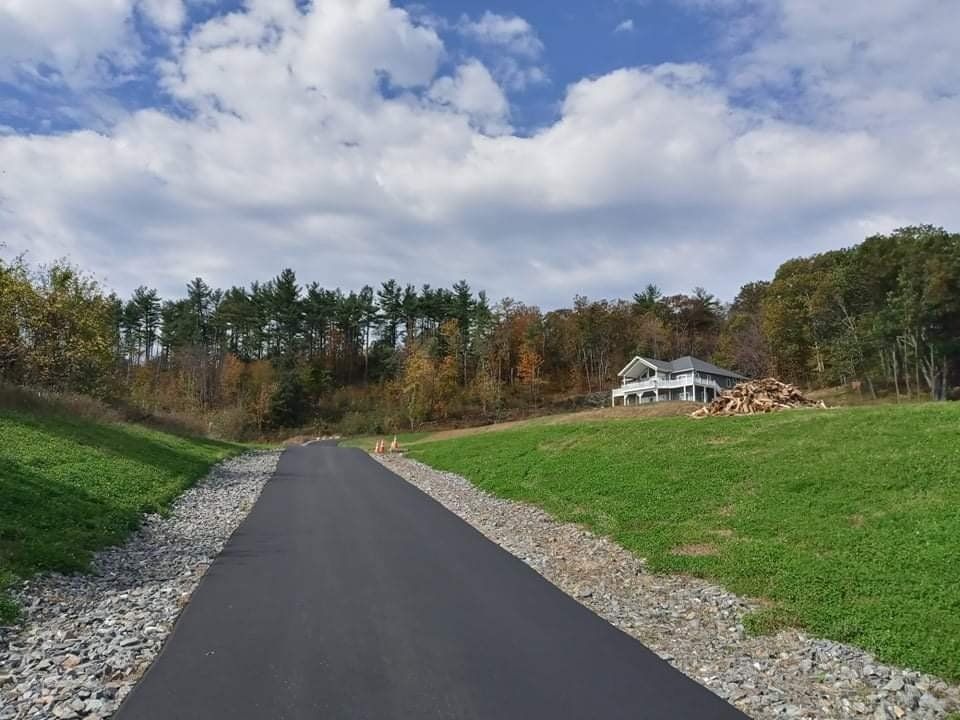 A paved road winds through a green, grassy slope toward a white house nestled among trees under a cloudy sky.