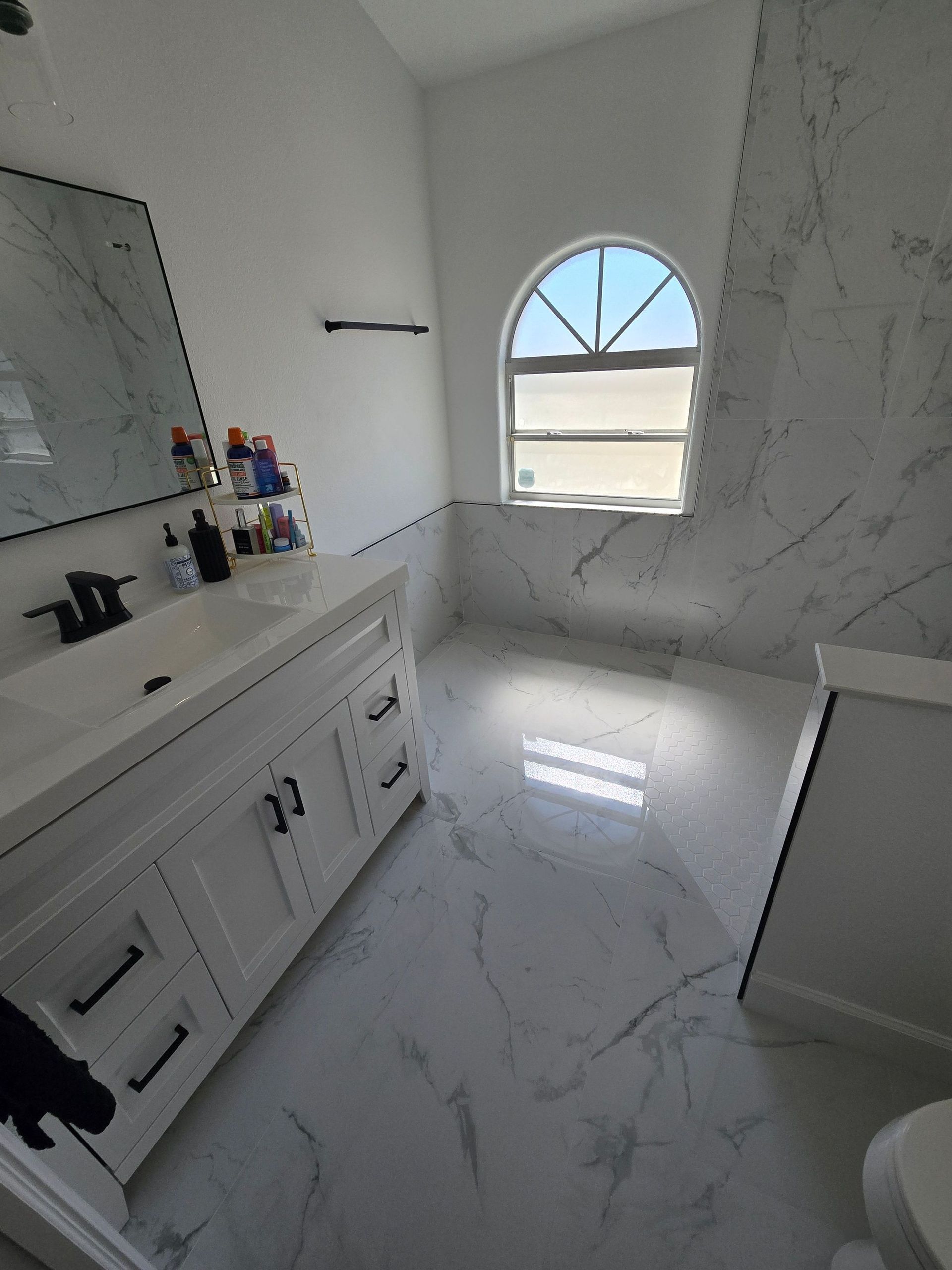 Bright modern bathroom with white marble tile, vanity, and arched window letting in sunlight.