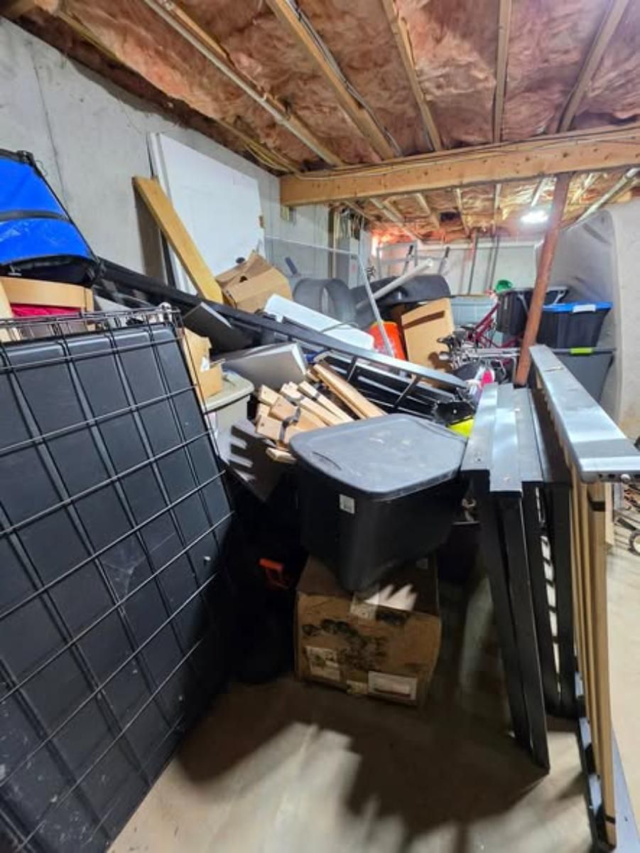 Cluttered basement or garage with stacked bins, tools, wood planks, and stored household items.