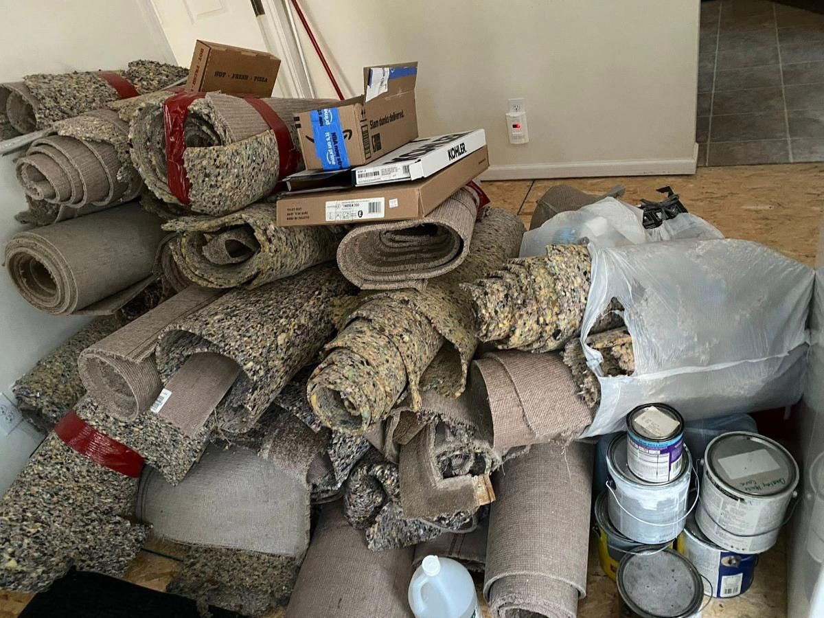 Pile of rolled carpets, cardboard boxes, and paint cans stacked in a room.