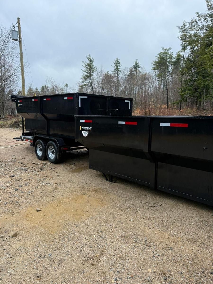Black dump trailer on gravel lot beside a black dumpster, with trees and utility poles in the background.