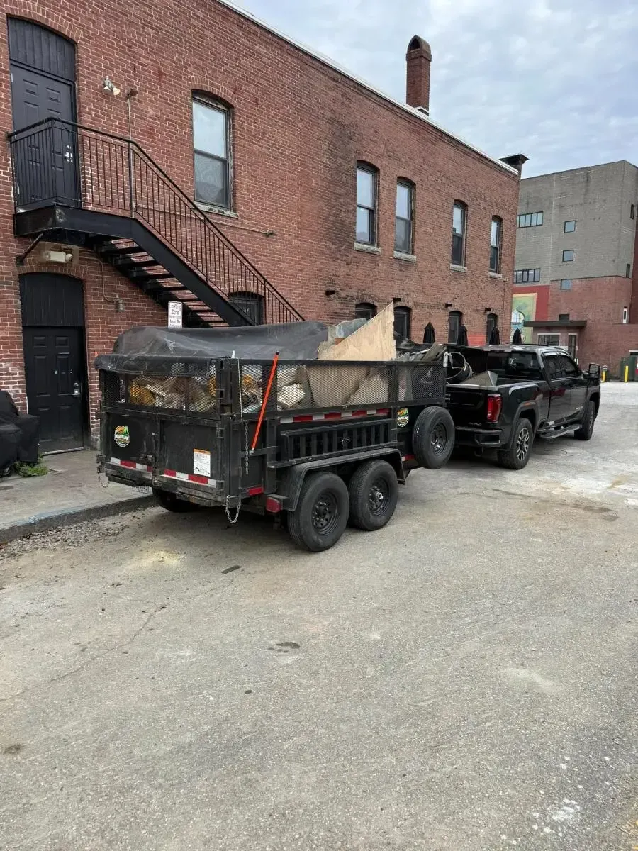 Black dump trailer and pickup truck parked beside a red brick building in an alley