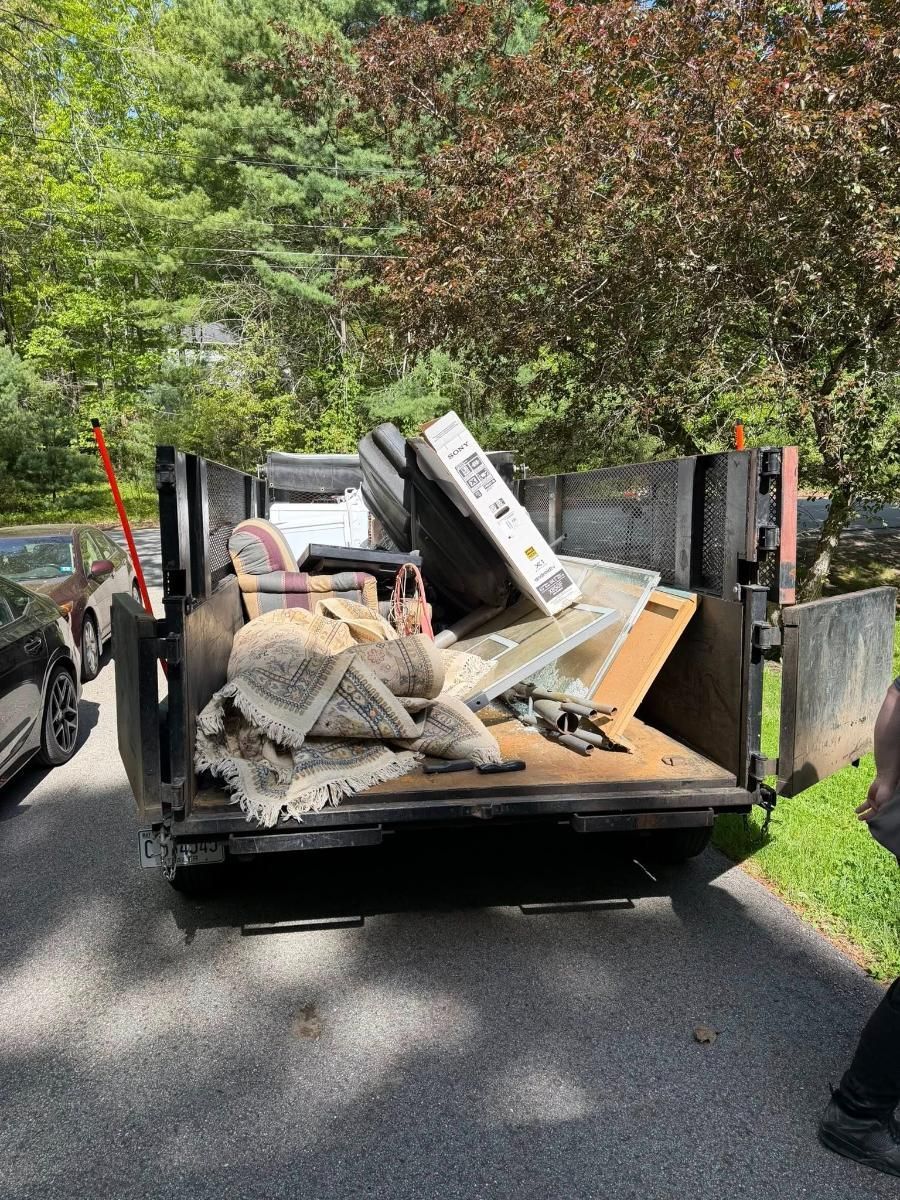 Loaded trailer with household debris and a mattress on a tree-lined street
