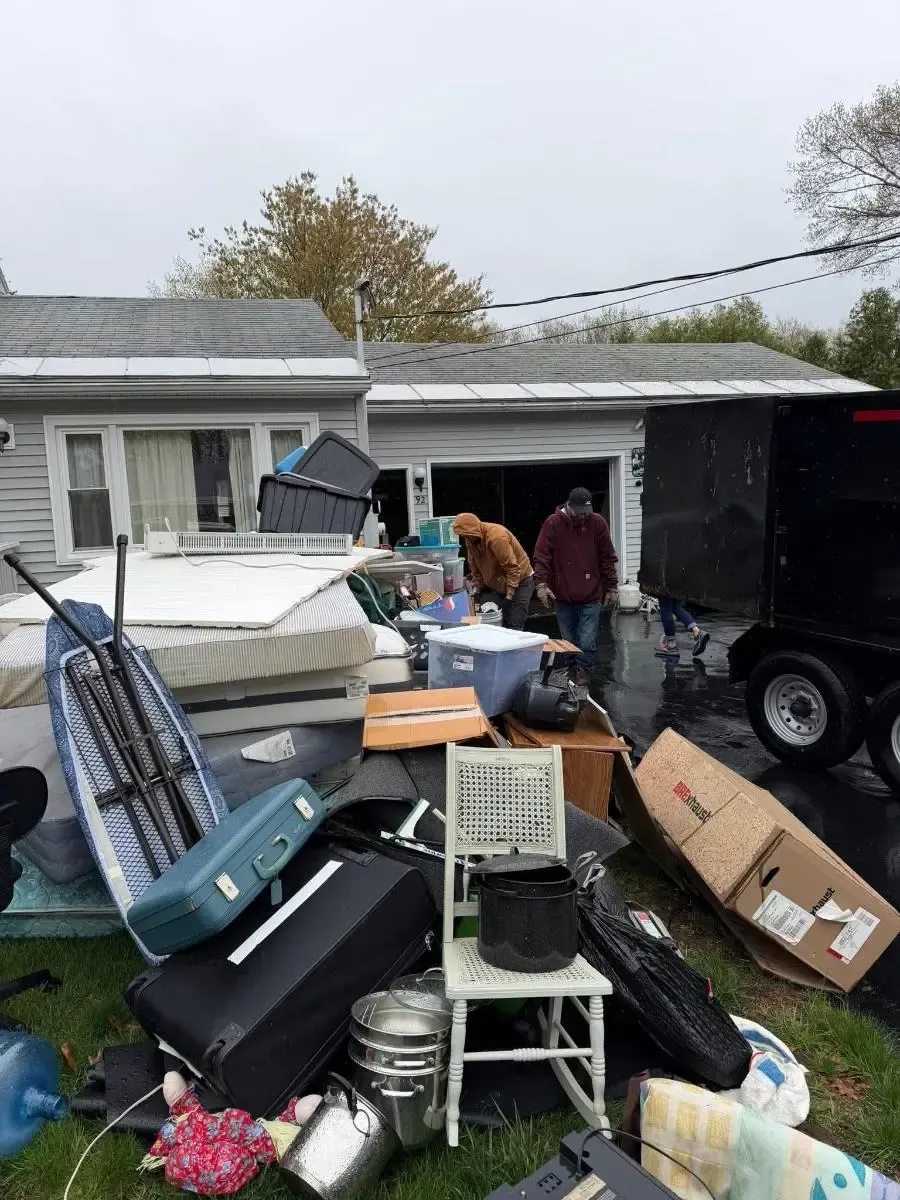 Cluttered driveway with piled furniture and boxes in front of a house, with two people standing nearby