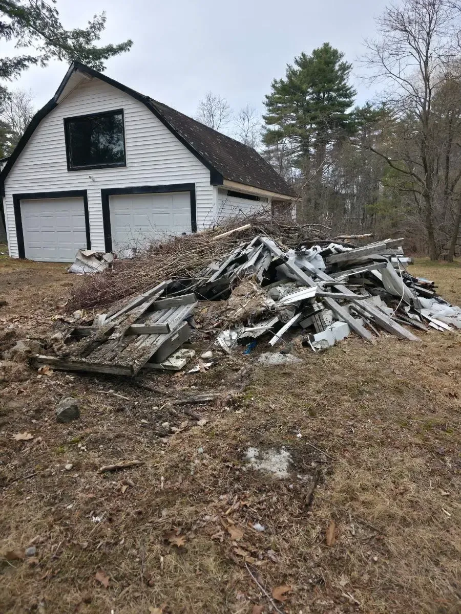 White house with a damaged roof beside a large pile of debris in a wooded yard