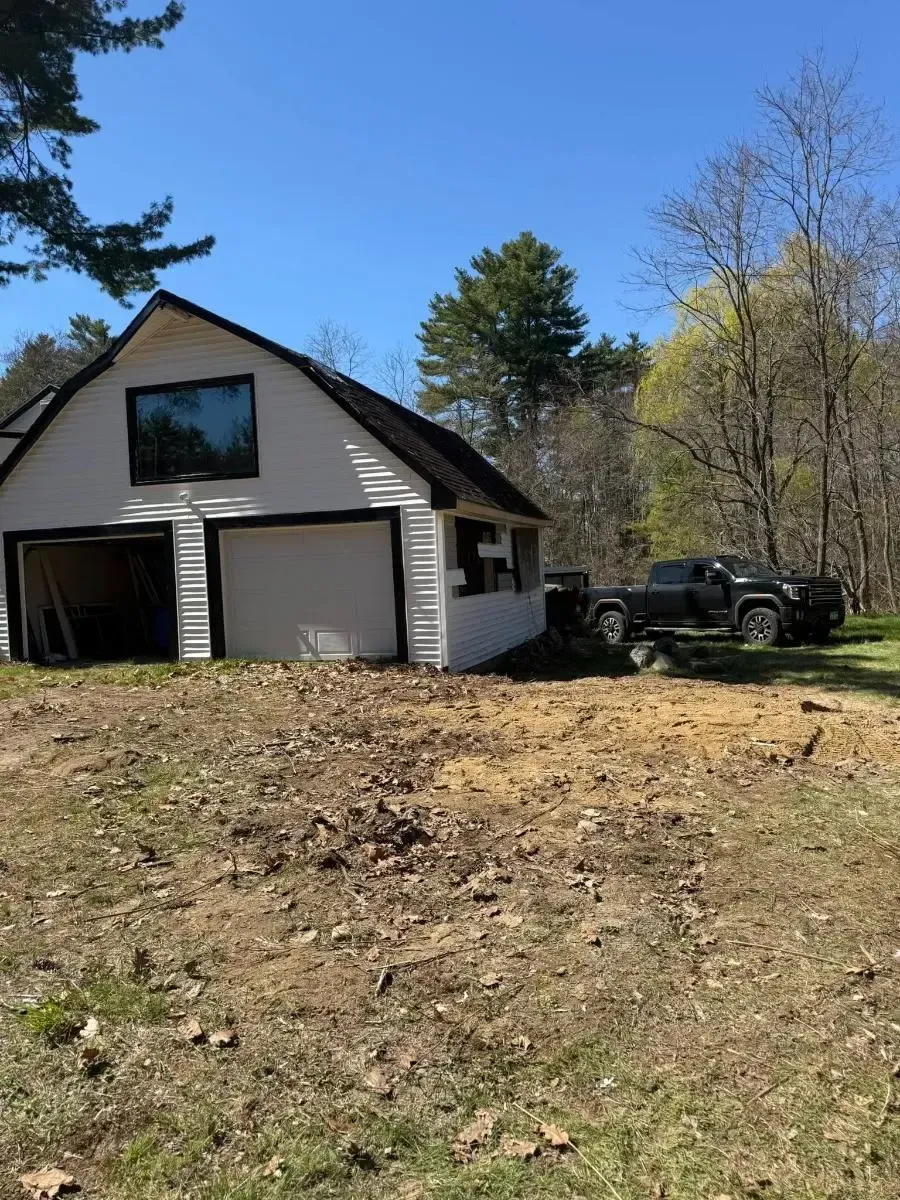 A white house with a garage sits on a grassy slope beside trees and a parked black pickup truck.