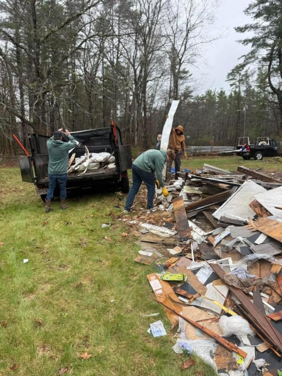 People cleaning debris and scrap wood into a dumpster in a muddy wooded yard
