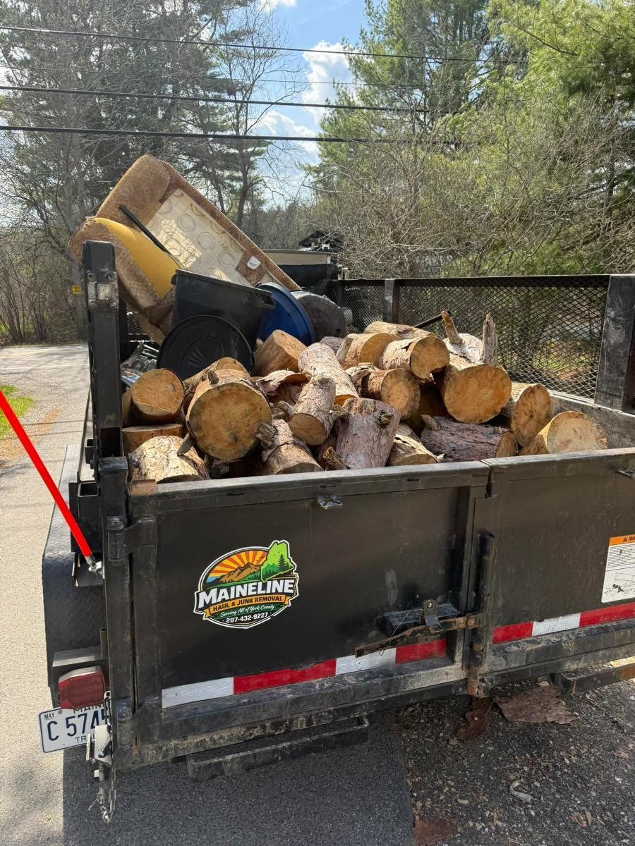 Dump trailer loaded with firewood on a gravel driveway near trees and a chain-link fence