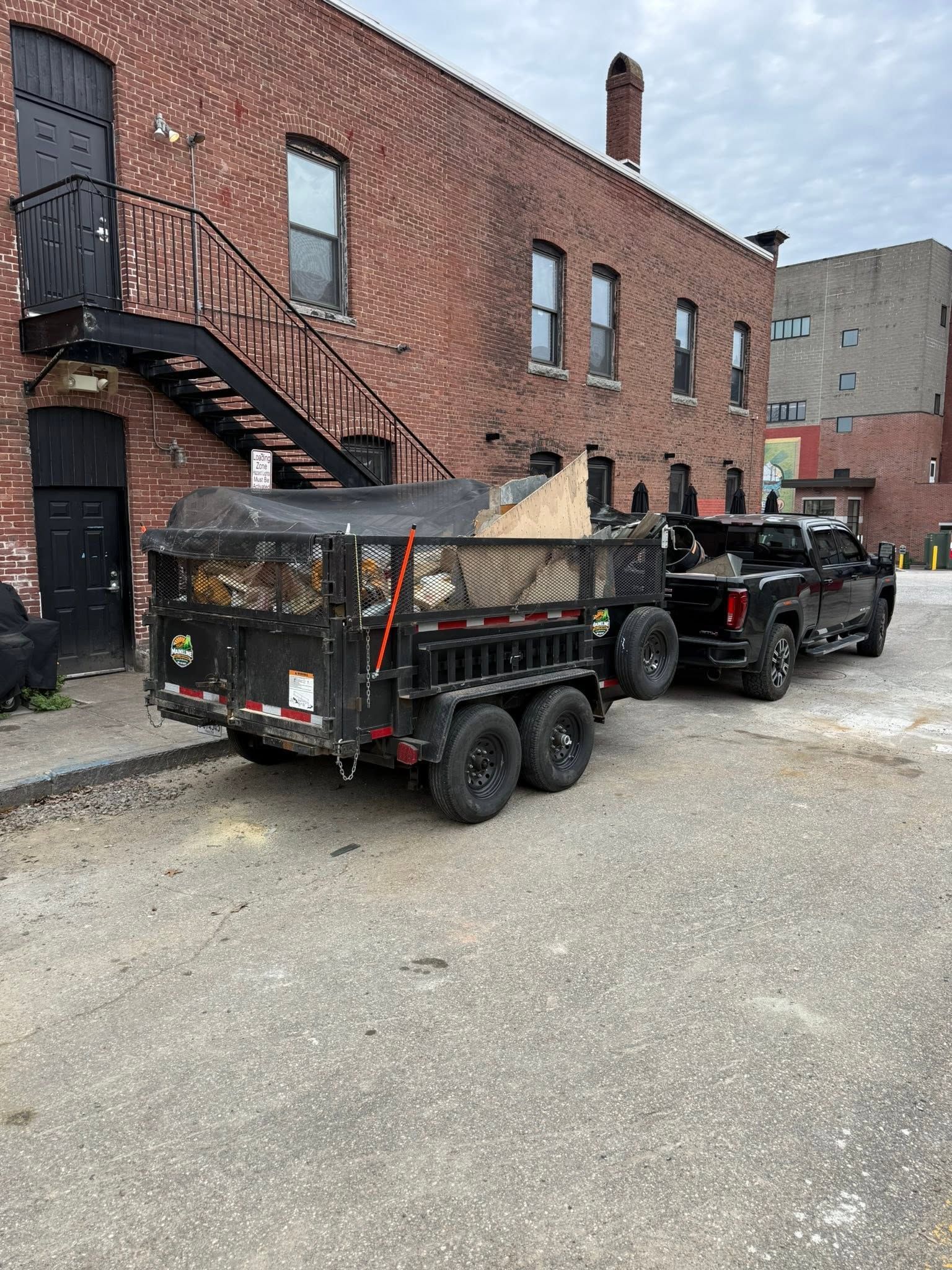 Black dump trailer hitched to a pickup in a brick alley beside a building with an exterior staircase