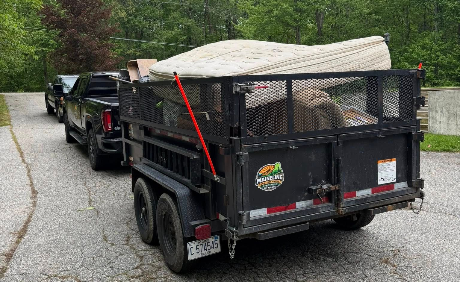 Black trailer loaded with gravel, hitched behind a pickup on a wooded gravel road.