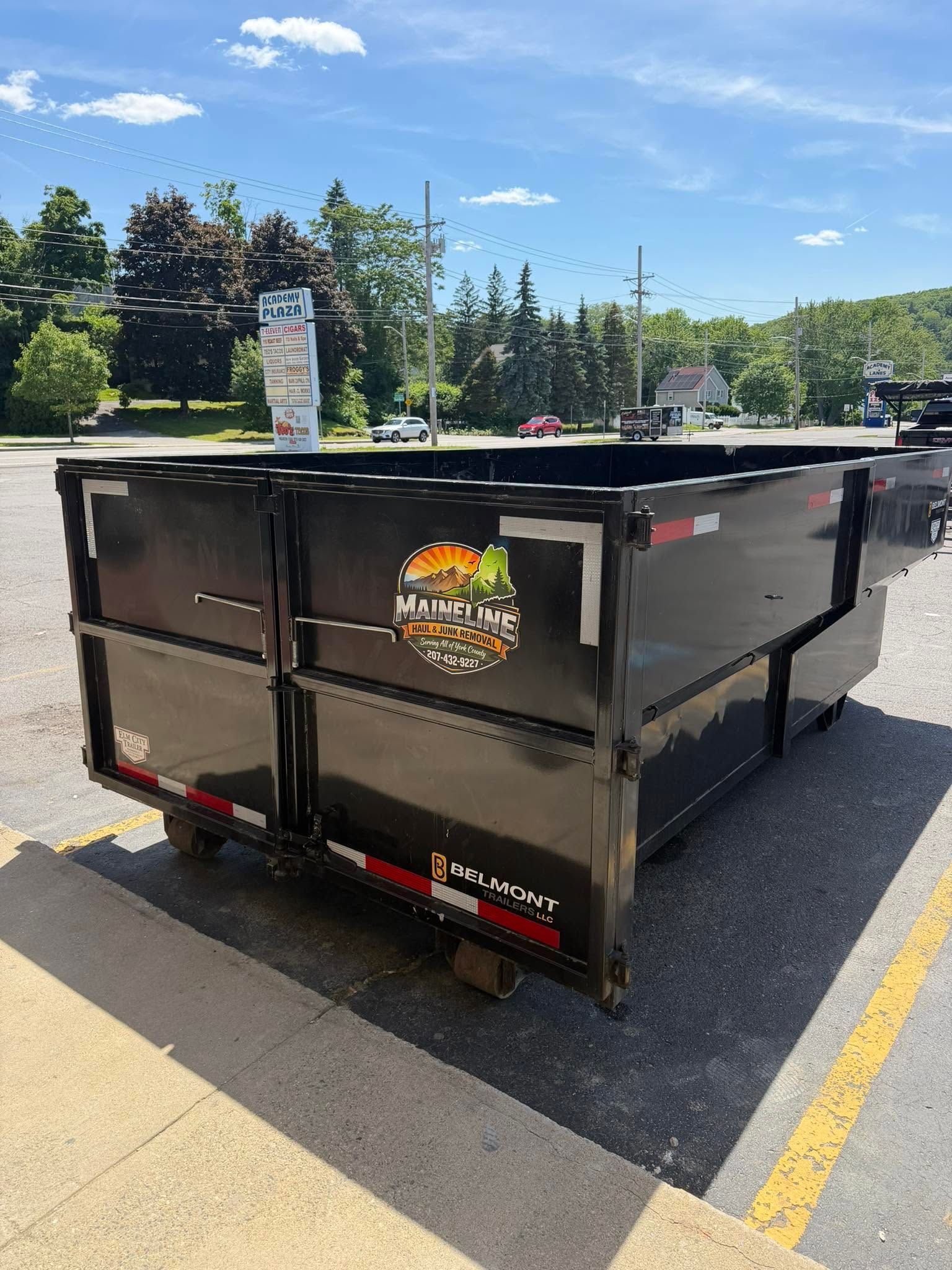 Black construction dumpster in a parking lot under a blue sky, with trees and road signs behind it
