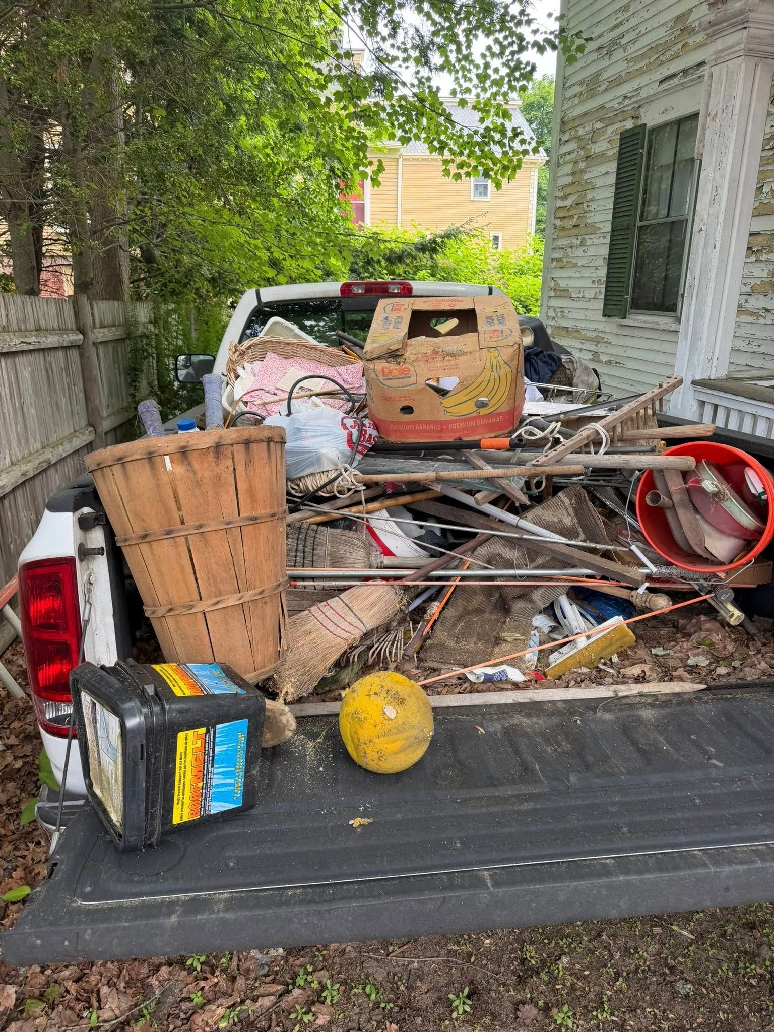 Outdoor trash pile on a truck bed, with assorted metal, baskets, and a yellow ball beside a house.