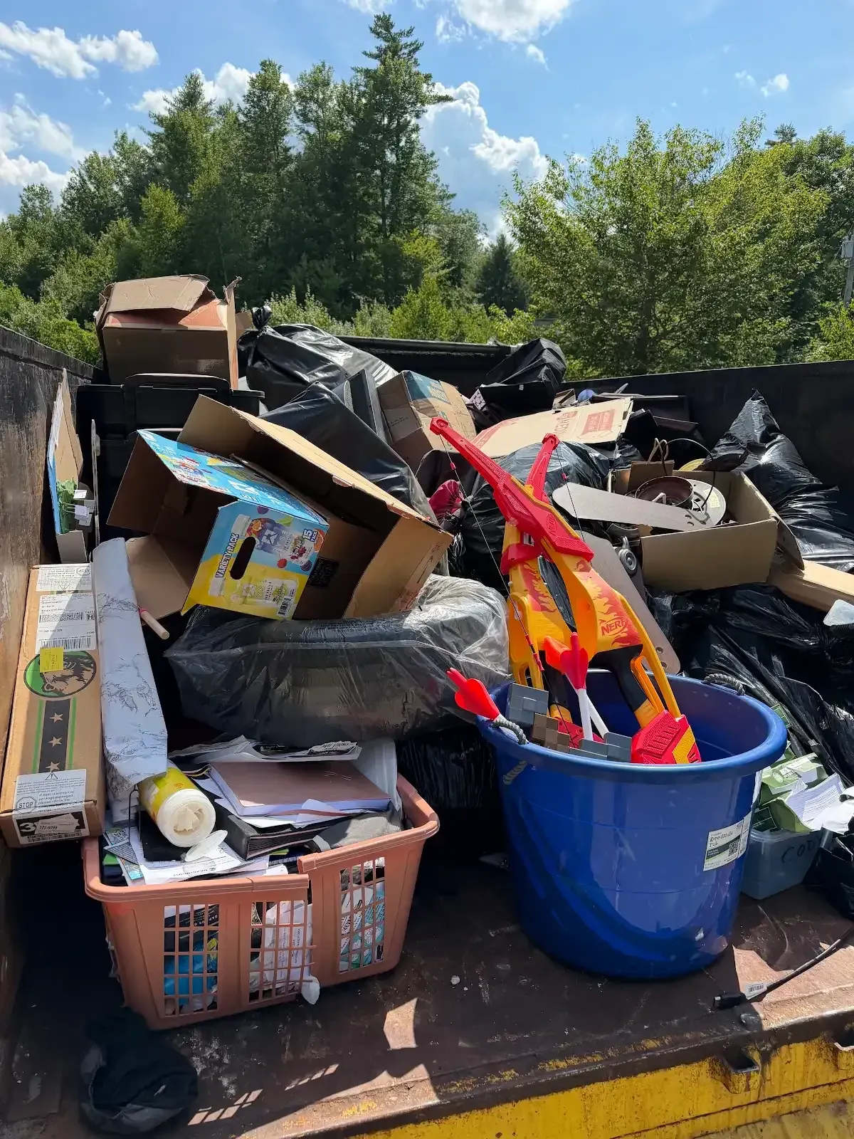 Overflowing trash bins and scattered cardboard in an outdoor waste area under trees.