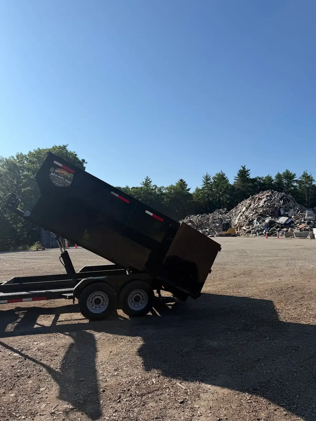 Black dump trailer raised on a gravel lot under a clear blue sky, with trees and debris piles in the background