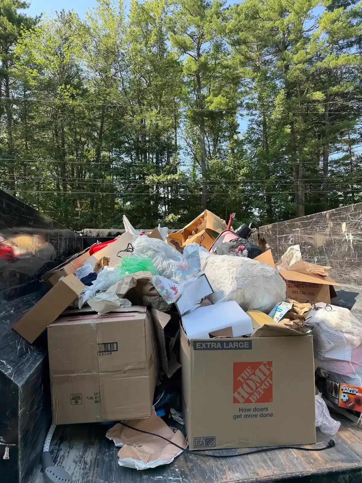 Overflowing dumpster with cardboard and trash bags in a wooded outdoor area