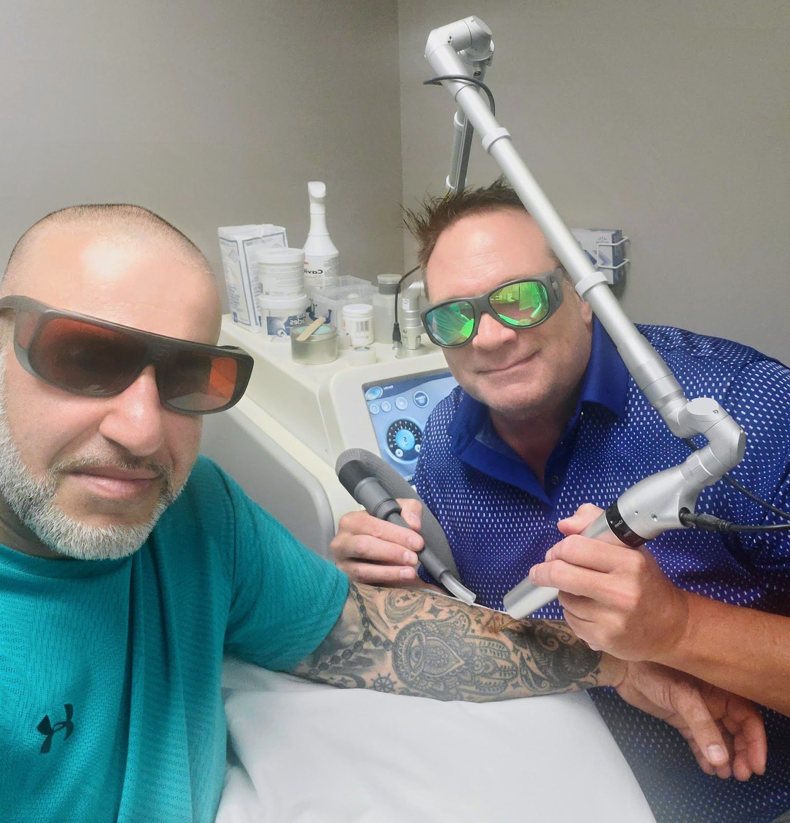Two men in a dental clinic take a selfie, one in an exam chair with a dental arm beside him.