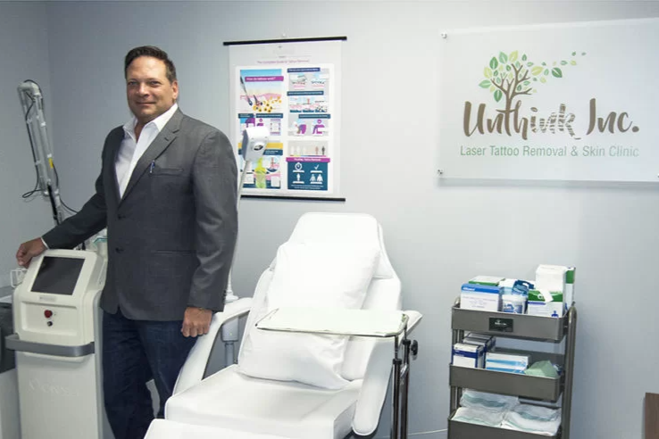 Man in a suit standing beside a medical treatment chair in a clinic office with equipment and branding on the wall