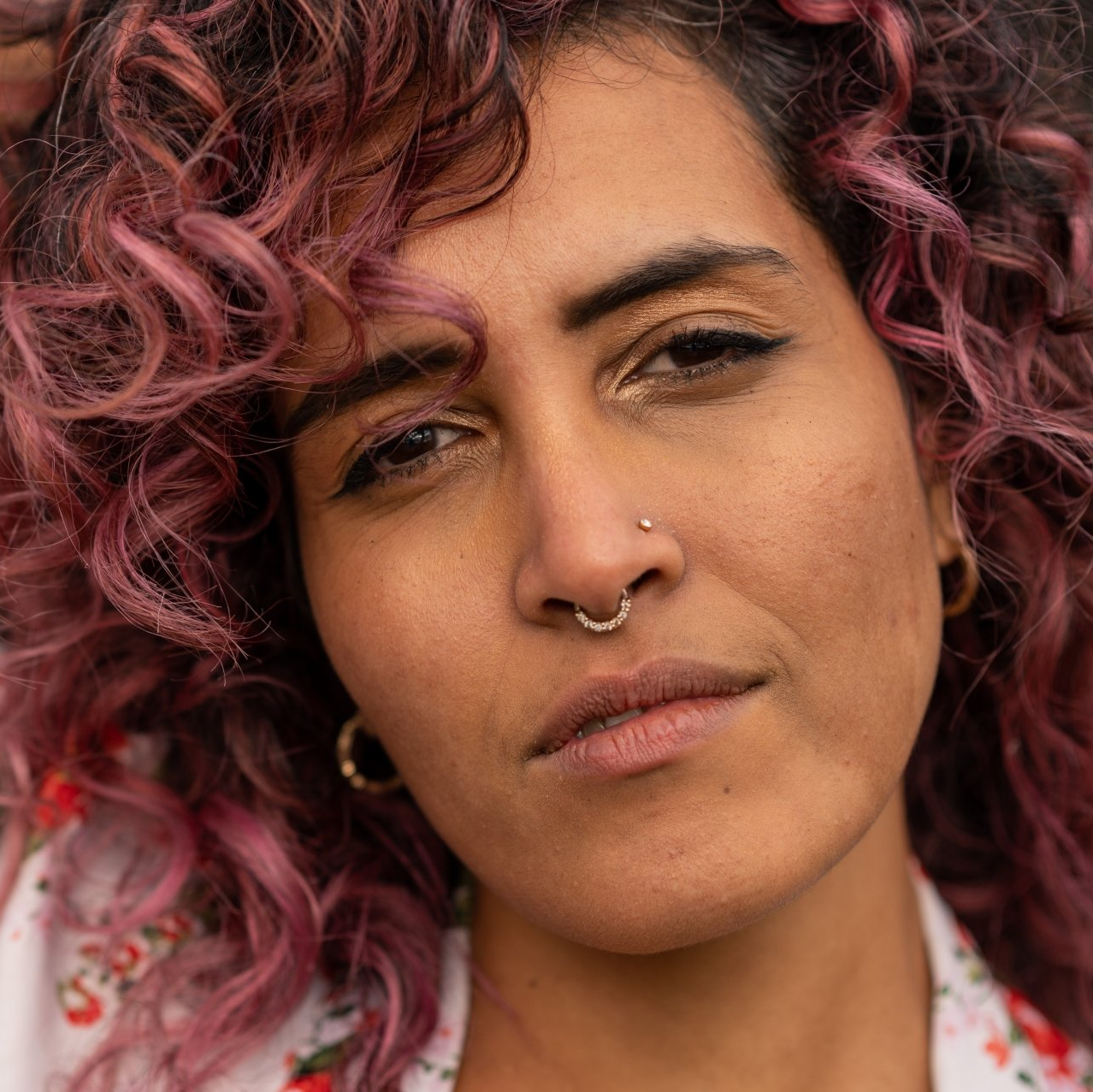 Close-up portrait of a person with curly pink hair, nose ring, and a neutral expression