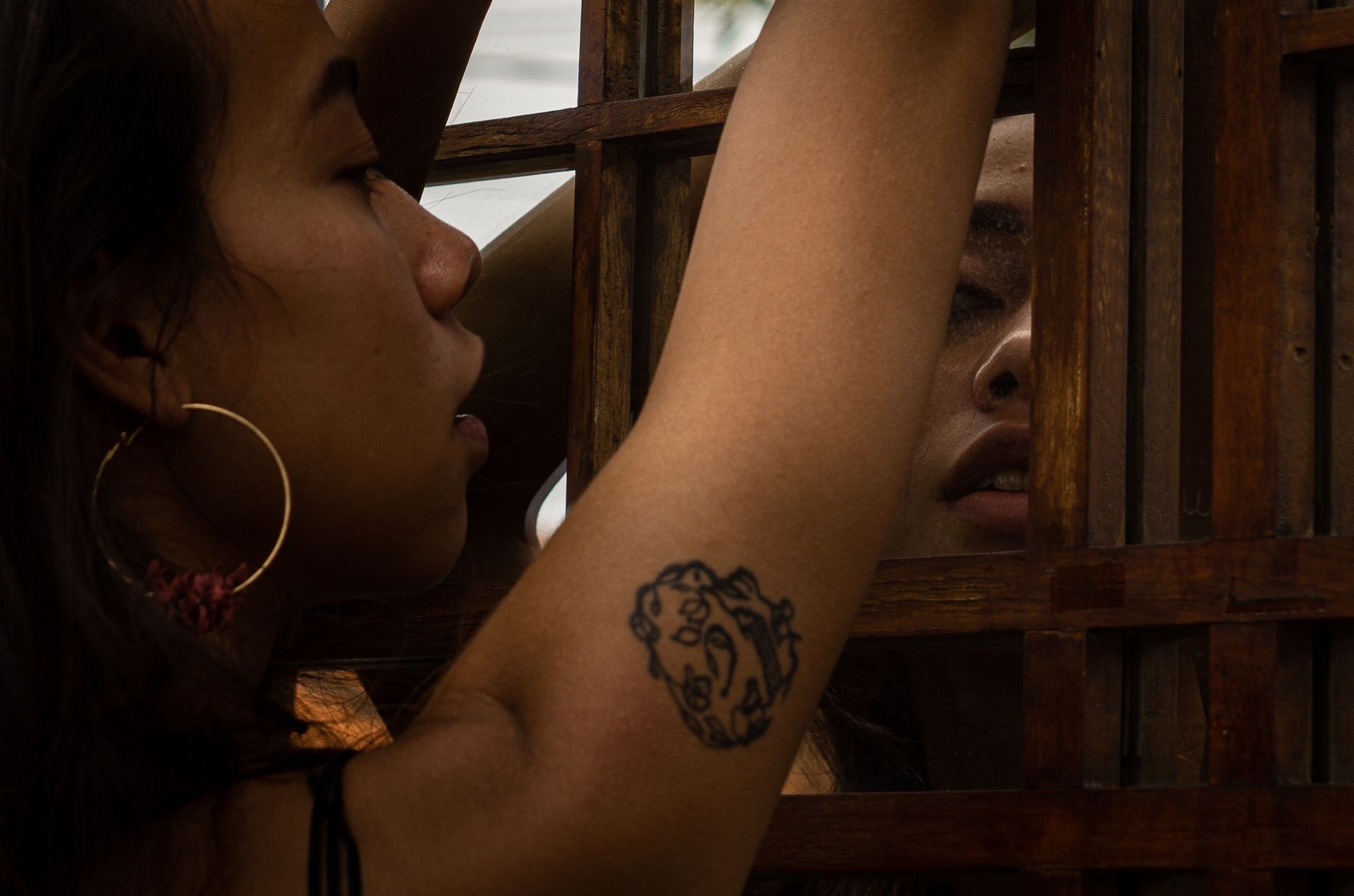 Woman with large hoop earring reaches toward a wooden window, with her reflection visible in the glass.