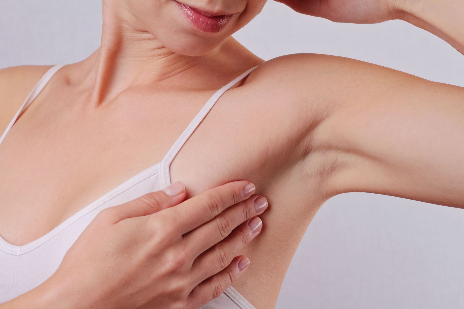 Woman examining her underarm in a white tank top against a light background